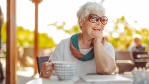 A senior women wearing sun glasses, smiling and holding a large ceramic coffee mug while sitting at a patio table in a wicker chair.
